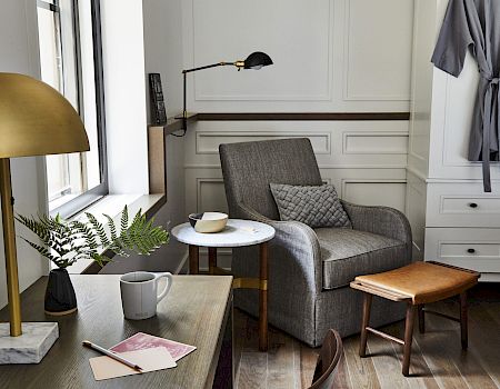 A cozy room features a gray armchair with a pillow, a table with a cup, notepad, lamp, and a robe hanging beside a cabinet.