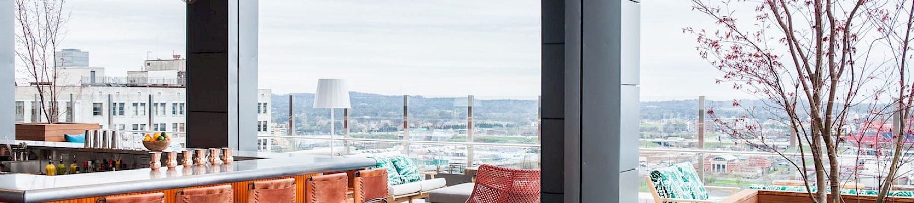 A rooftop bar with modern seating, colorful cushions, and a city view, featuring trees and a stylish counter with bar stools.