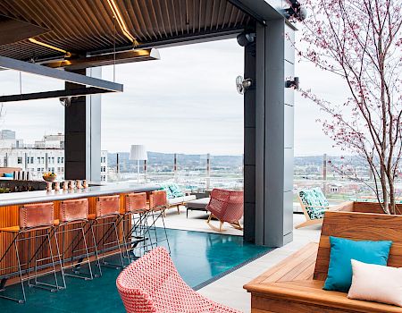 A rooftop bar with modern seating, colorful cushions, and a city view, featuring trees and a stylish counter with bar stools.