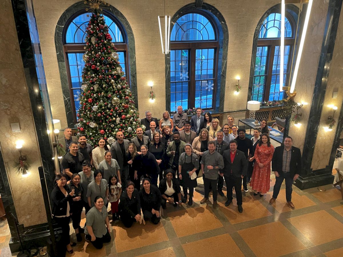 A group of people pose together in a festive setting with a large Christmas tree inside a decorated room with arched windows and warm lighting.