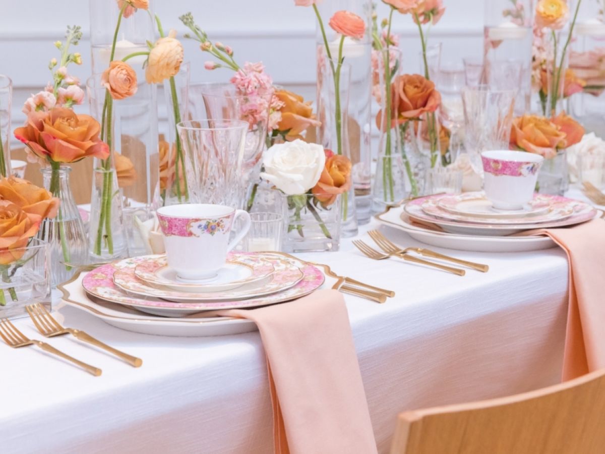 Elegant table setting with floral plates, teacups, peach napkins, gold cutlery, and orange-pink flowers in glass vases.