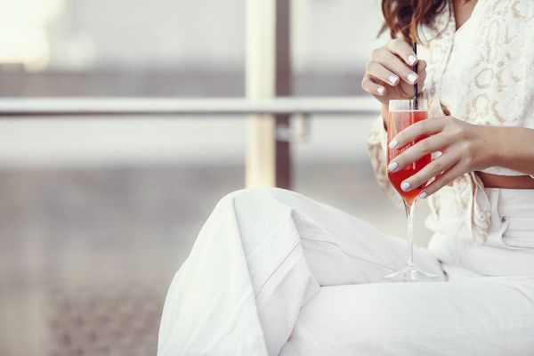 A person in white clothing holds a red drink in a flute glass, stirring it with a straw, sitting in a relaxed outdoor setting.