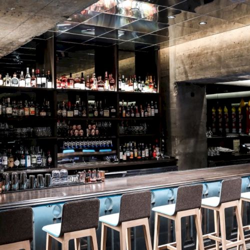 A modern bar with a long counter, high chairs, and a well-stocked shelf of bottles against a concrete backdrop, featuring ambient lighting.