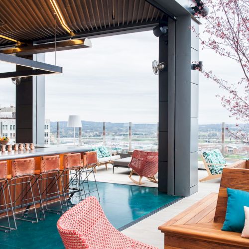 A stylish rooftop bar with bar stools, modern seating, and decorative plants, offering panoramic views of the cityscape under a cloudy sky.