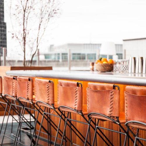 A rooftop bar features leather-backed chairs, a counter with a basket of oranges, metal shakers, and a cityscape view in the background.