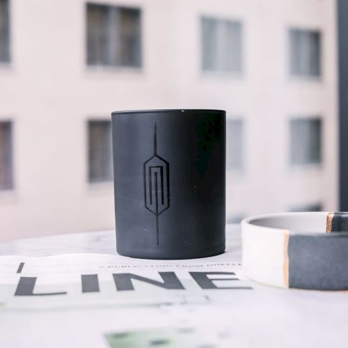 A black candle holder with a geometric design and a ceramic bowl sit on a magazine by a window with an out-of-focus building view.