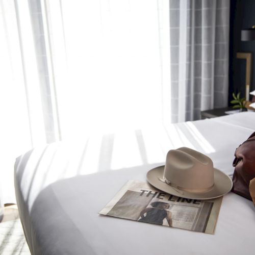 The image shows a bed with a hat, newspaper, and a bag on it, with sunlight streaming through sheer curtains.