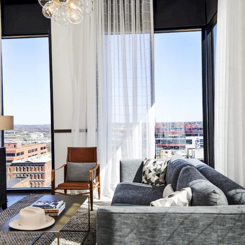 A modern living room with a gray couch, a chair, large windows, curtains, a rug, a TV, and a city view in the background.