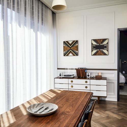 A modern dining room with a wooden table, decorative plate, and sideboard in front of two abstract artworks, beside sheer curtains.