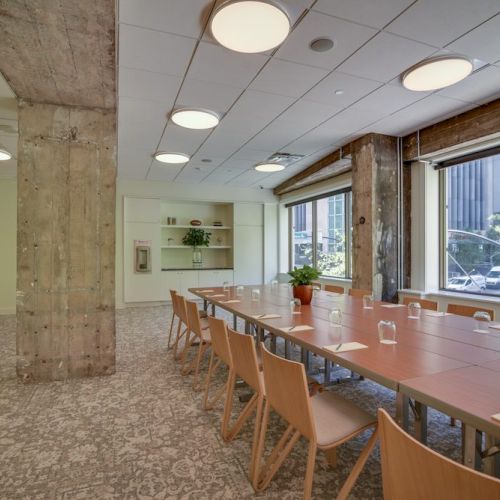 A modern conference room with a long table, chairs, concrete beams, large windows, and bright lighting, viewed with a carpeted floor.