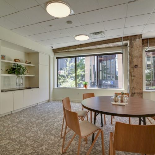 A modern office space with wooden chairs around a table, large window views, shelving, and a wall-mounted coffee machine.