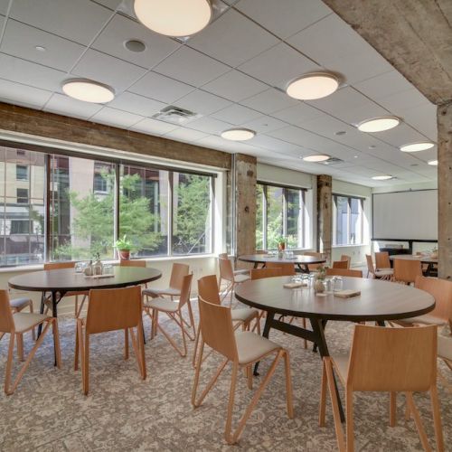 A modern dining area with wooden tables and chairs, large windows providing natural light, and exposed concrete columns.