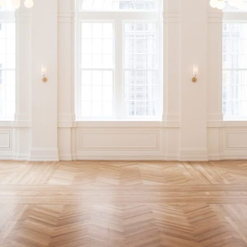 A bright, empty room with wooden herringbone floors, large windows, and white walls featuring elegant sconces.