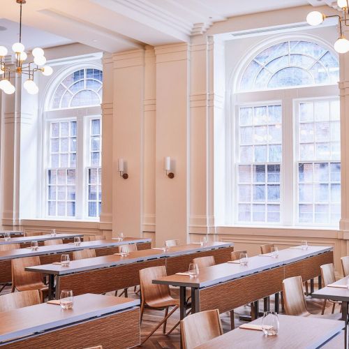 The image shows a bright, elegant conference room with rows of tables, chairs, large arched windows, and modern chandeliers.