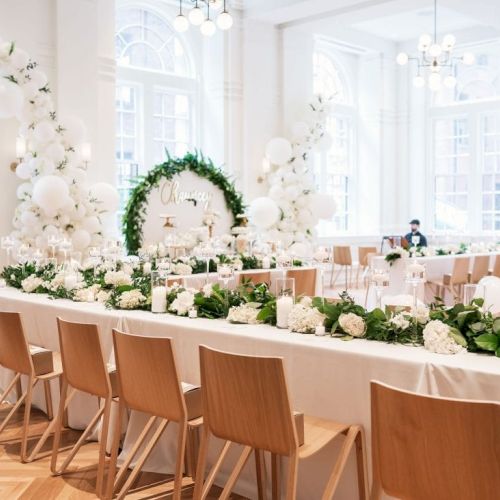 A beautifully decorated event space with white flowers, balloons, wooden chairs, and greenery on tables, set in a bright, elegant room.