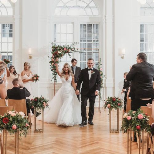 A bride and groom walk down the aisle, surrounded by guests in a bright, elegant room decorated with flowers and wooden chairs.