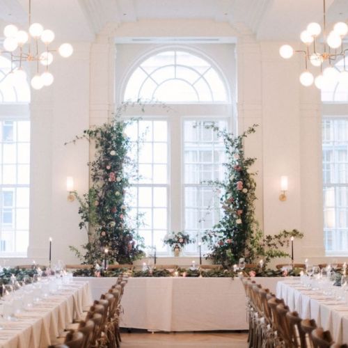 The image shows an elegantly set banquet hall with long tables, white tablecloths, floral centerpieces, and large arched windows.