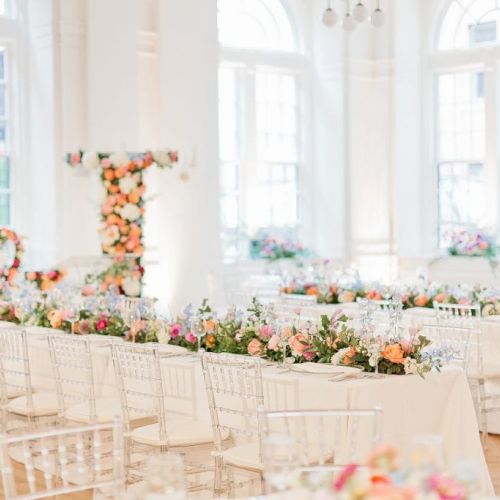 Elegant wedding reception with floral decorations, white chairs, and large windows filling the space with natural light.