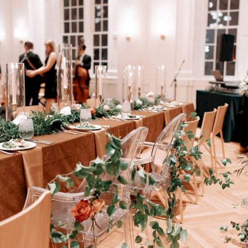 Elegantly decorated event space with a long table, green foliage, candles, and transparent chairs; people stand near the back.