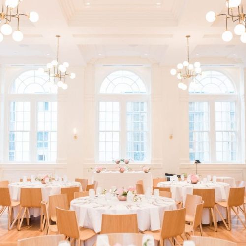 A bright, elegant dining room with round tables, white tablecloths, wooden chairs, and decorative lights, ready for an event or gathering.