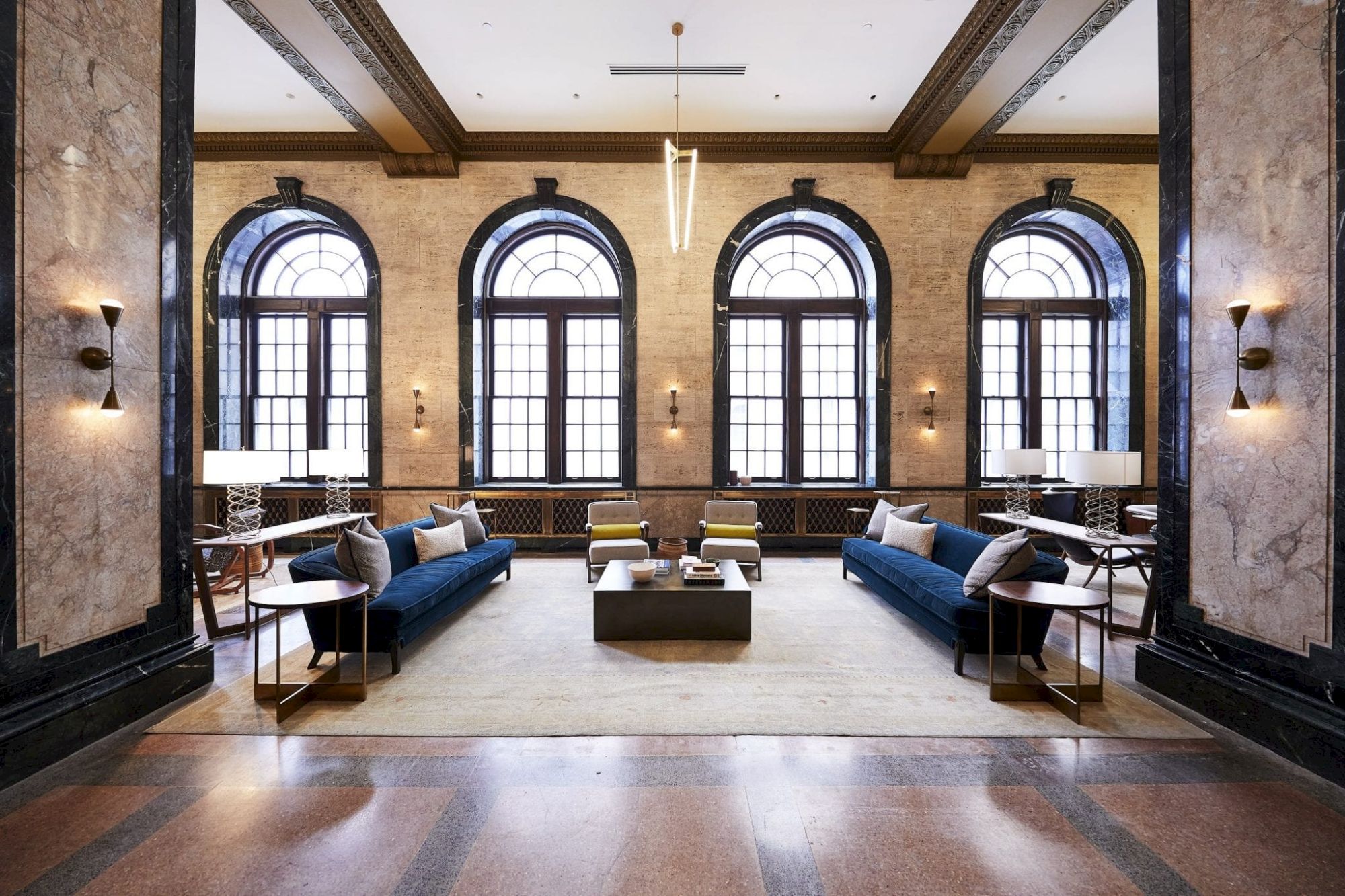 A spacious, elegant room features large arched windows, two blue sofas, chairs, a central coffee table, and decorative lighting.