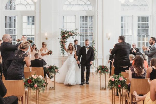 A newlywed couple walks down the aisle in a bright, elegant venue, with guests clapping and celebrating their union.