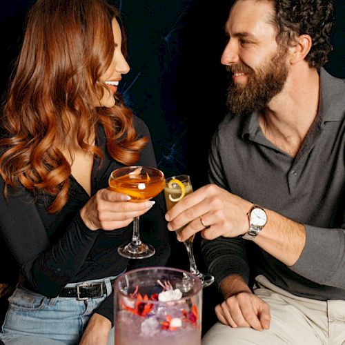 A smiling couple in casual attire clinks cocktails over a table with a festive ice bucket.