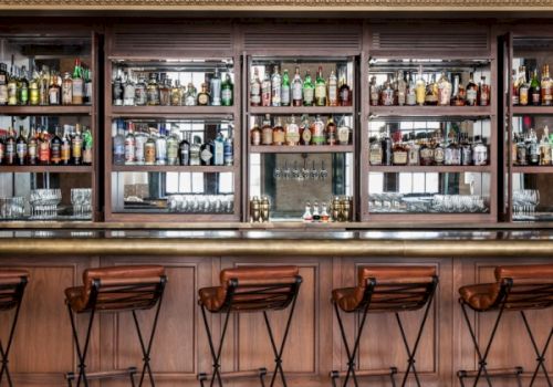 A well-stocked bar with wooden stools lined up in front of shelves filled with various bottles and glasses, creating a cozy atmosphere.