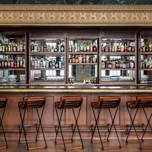 A well-stocked bar with wooden stools lined up in front of shelves filled with various bottles and glasses, creating a cozy atmosphere.