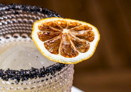 A cocktail in a textured glass with a black salt rim, garnished with a slice of dried citrus fruit on the edge.