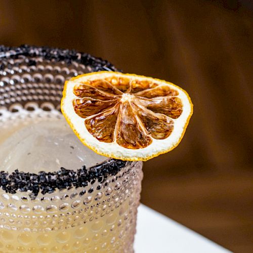 A cocktail in a textured glass with a black salt rim, garnished with a slice of dried citrus fruit on the edge.