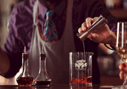 A bartender is mixing a drink, using a jigger to pour into a beaker. Two bottles with liquid are on the counter.