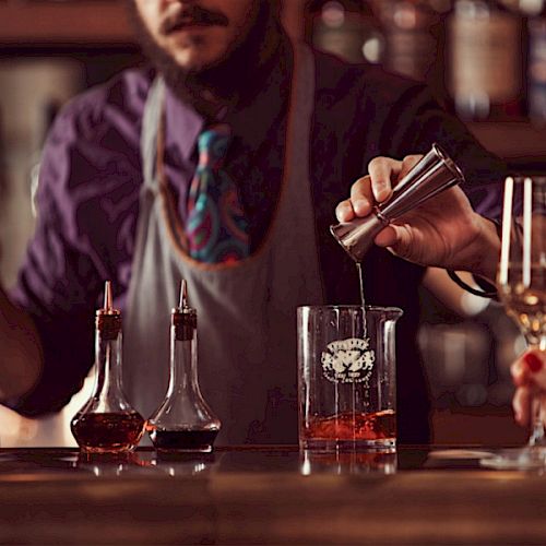 A bartender is mixing a drink, using a jigger to pour into a beaker. Two bottles with liquid are on the counter.