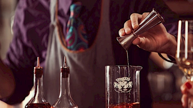 A bartender is mixing a drink, using a jigger to pour into a beaker. Two bottles with liquid are on the counter.