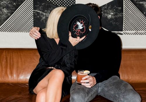 A couple on a couch covers their faces with a black hat. The woman wears black and holds a drink, and there's abstract art on the wall.