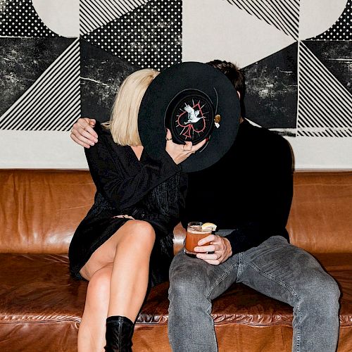 A couple on a couch covers their faces with a black hat. The woman wears black and holds a drink, and there's abstract art on the wall.