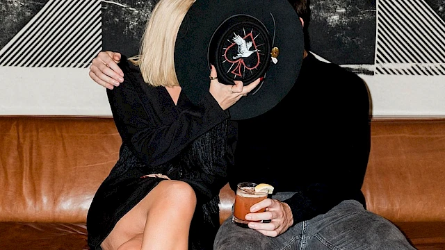 A couple on a couch covers their faces with a black hat. The woman wears black and holds a drink, and there's abstract art on the wall.