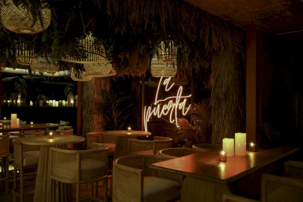 A cozy, dimly lit restaurant with candles on wooden tables, thatched decor, and a neon sign reading 