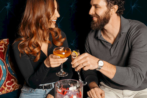 A man and woman are smiling and toasting with cocktails. There's a decorative cushion and a beverage bucket with flowers nearby.