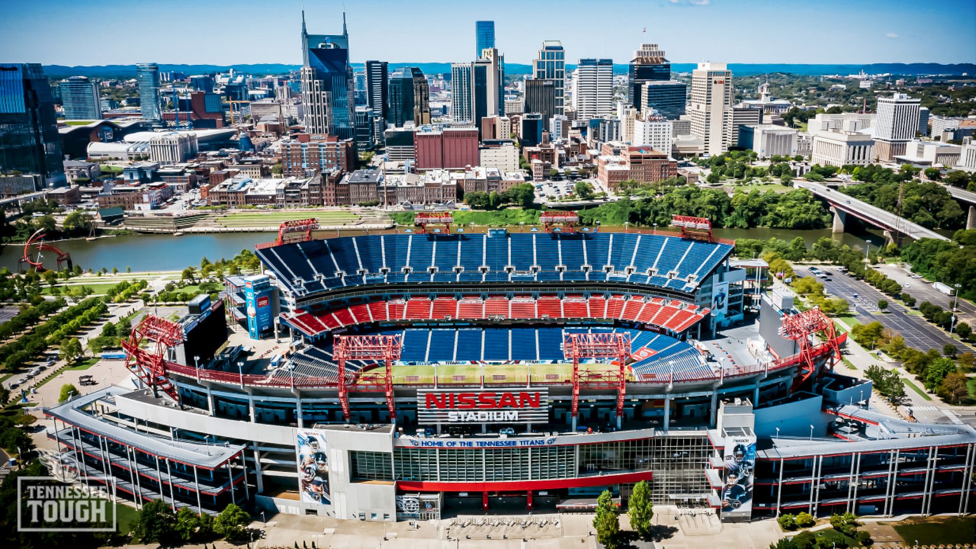 An aerial view of a large stadium with a city skyline in the background, surrounded by green space and water.