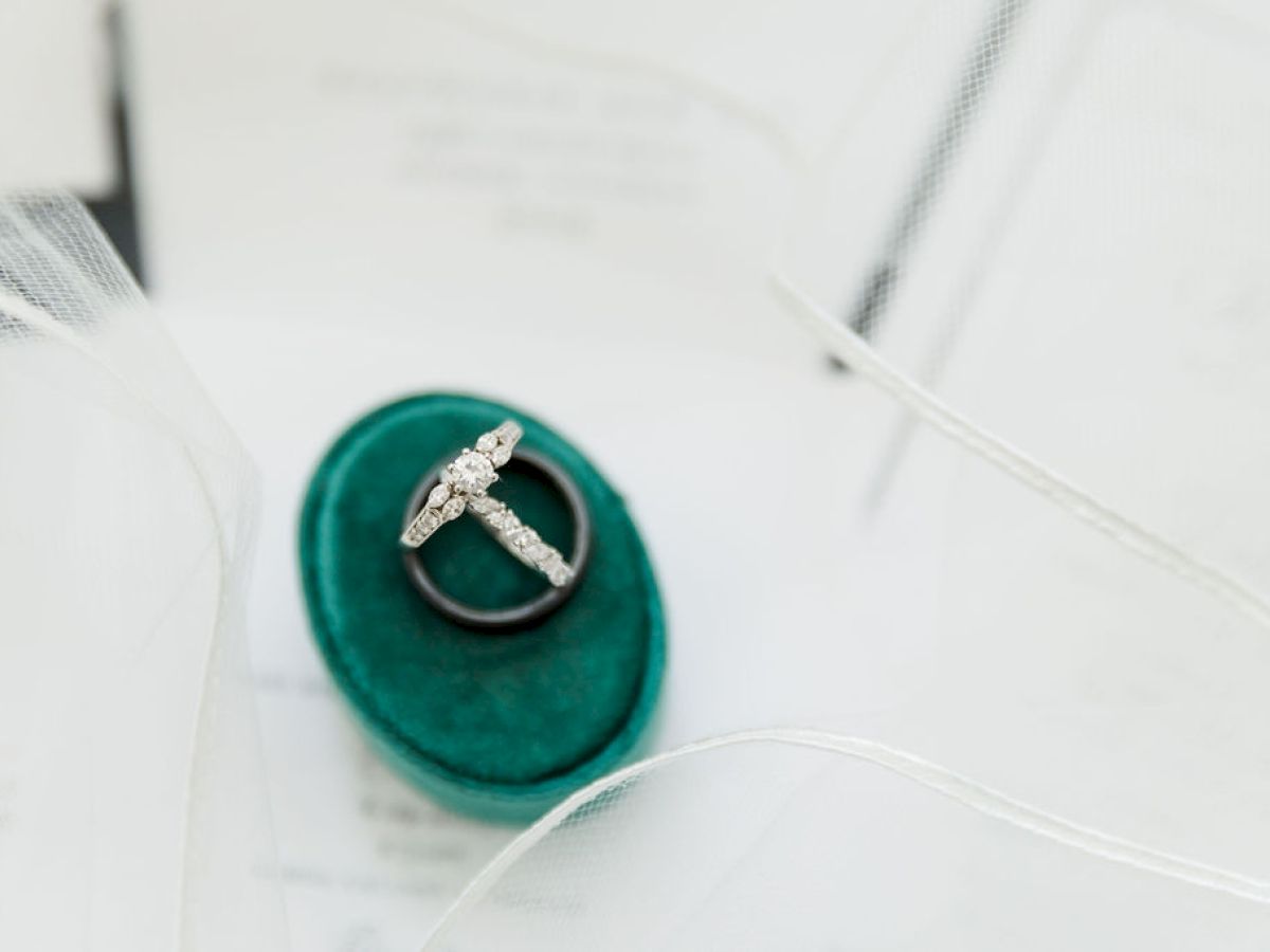 A diamond ring set is displayed on a teal velvet box, with tulle fabric and paper invitations in the background, capturing a bridal theme.