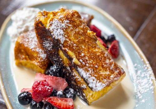 French toast with berries and powdered sugar on top, served with a creamy sauce on an oval plate.