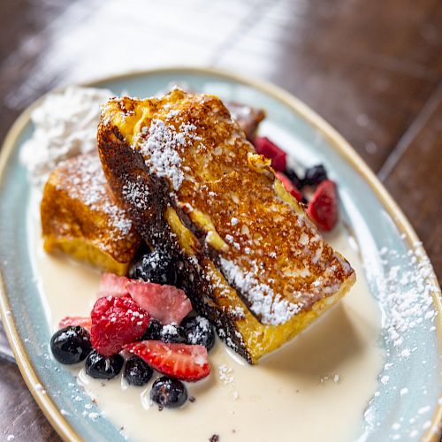 French toast with berries and powdered sugar on top, served with a creamy sauce on an oval plate.