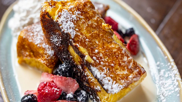 French toast with berries and powdered sugar on top, served with a creamy sauce on an oval plate.