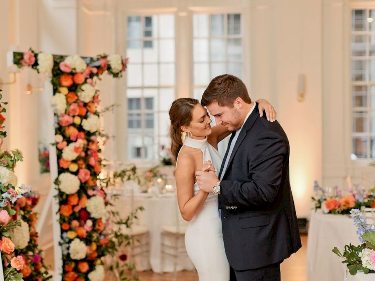 A couple dances together in a beautifully decorated room with flower arrangements. There are tables set with floral centerpieces.