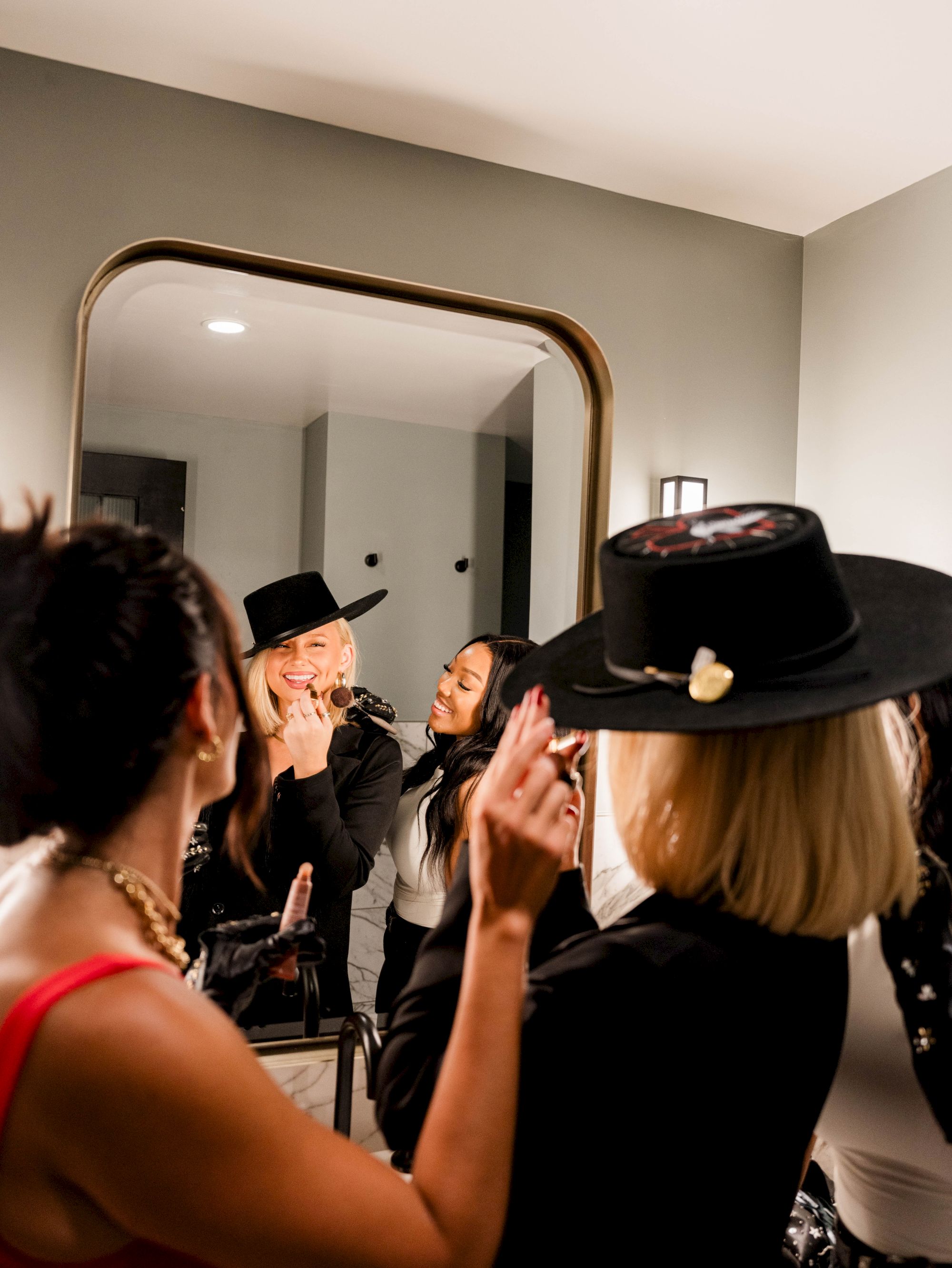 A group of people in front of a mirror, some wearing hats, appear to be getting ready or enjoying themselves in a well-lit room.