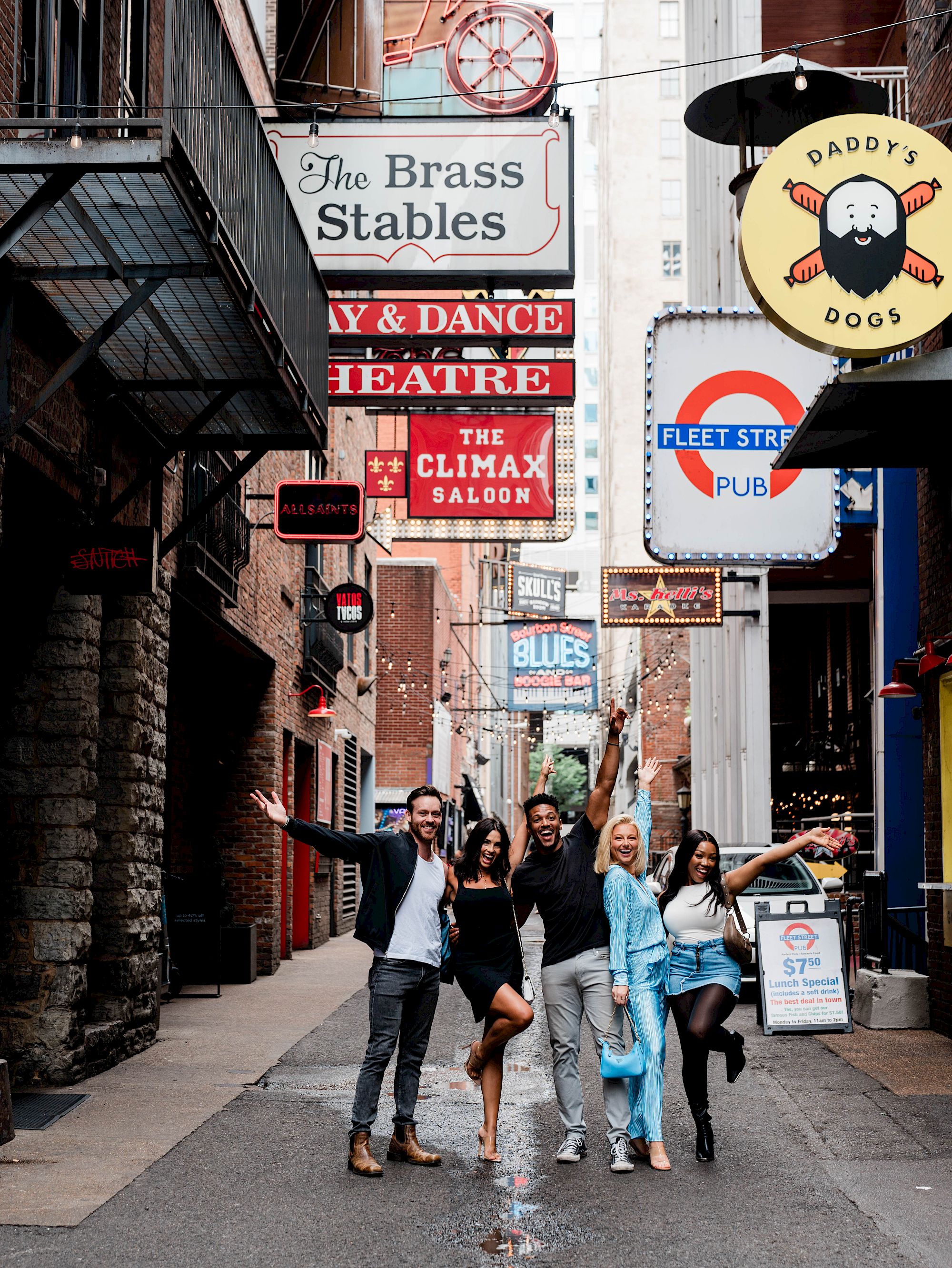 A group of five people are posing energetically in an alleyway lined with colorful signs for bars and theaters.