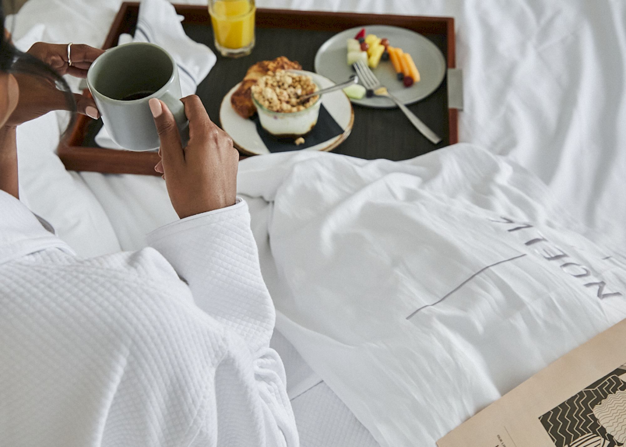 A person in a robe holds a mug on a bed with a breakfast tray, featuring fruit, cereal, and juice, next to a newspaper.