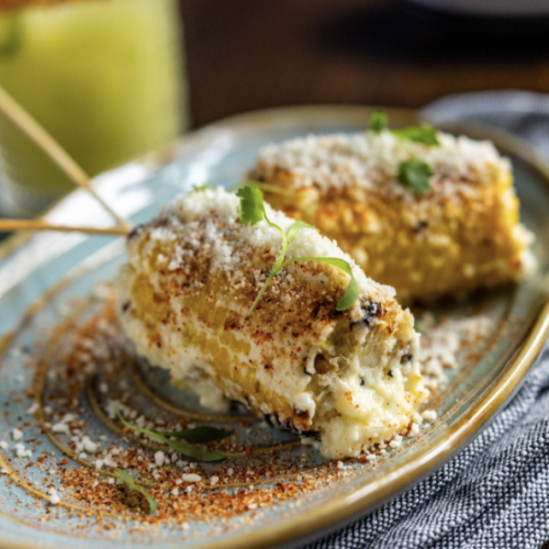 Grilled corn on the cob topped with cheese and spices on a plate, served on a textured cloth, with a drink in the background.