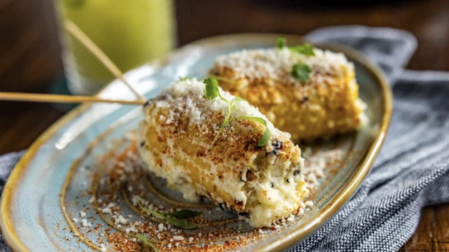 Grilled corn on the cob topped with cheese and spices on a plate, served on a textured cloth, with a drink in the background.